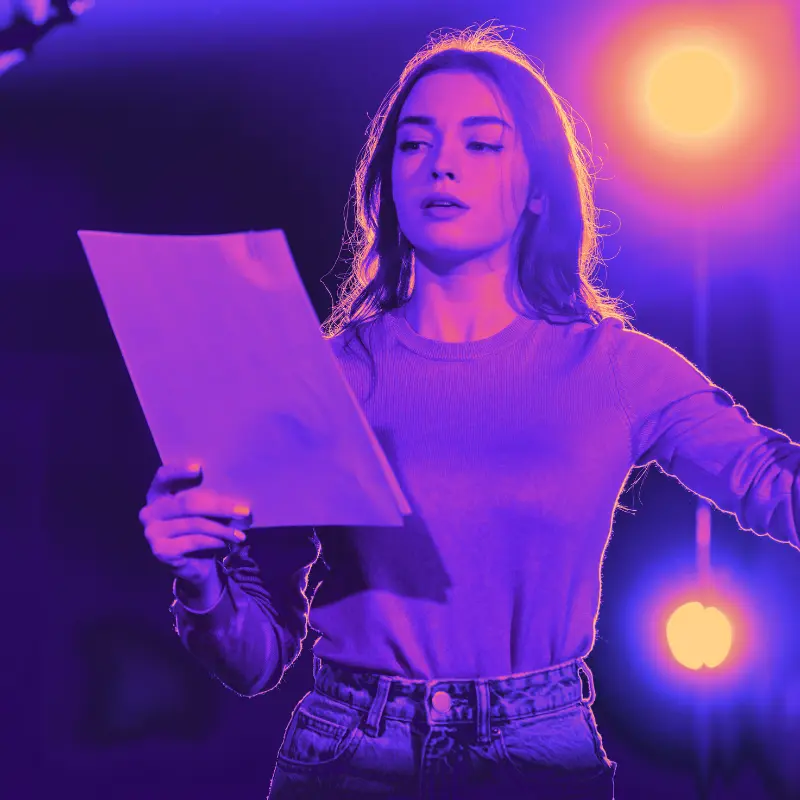 A woman stands on stage during theatre auditions, reading lines from a script with a bright spotlight behind her, casting a warm glow that highlights her focused expression.