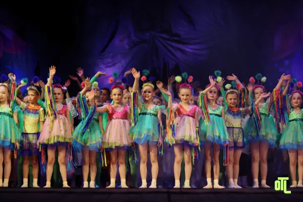 A group of young children in colorful costumes standing on stage, waving happily, representing youth involvement in theatre before main events.