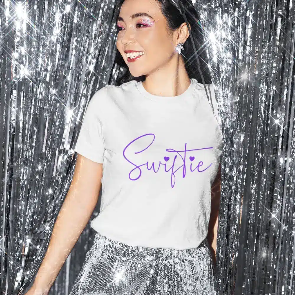 Smiling woman wearing a white t-shirt with the word "Swiftie" in purple script, standing in front of sparkling silver tinsel.