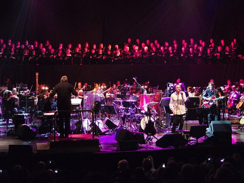 A large ensemble of singers and musicians performing under vivid stage lighting during Meltdown festival.