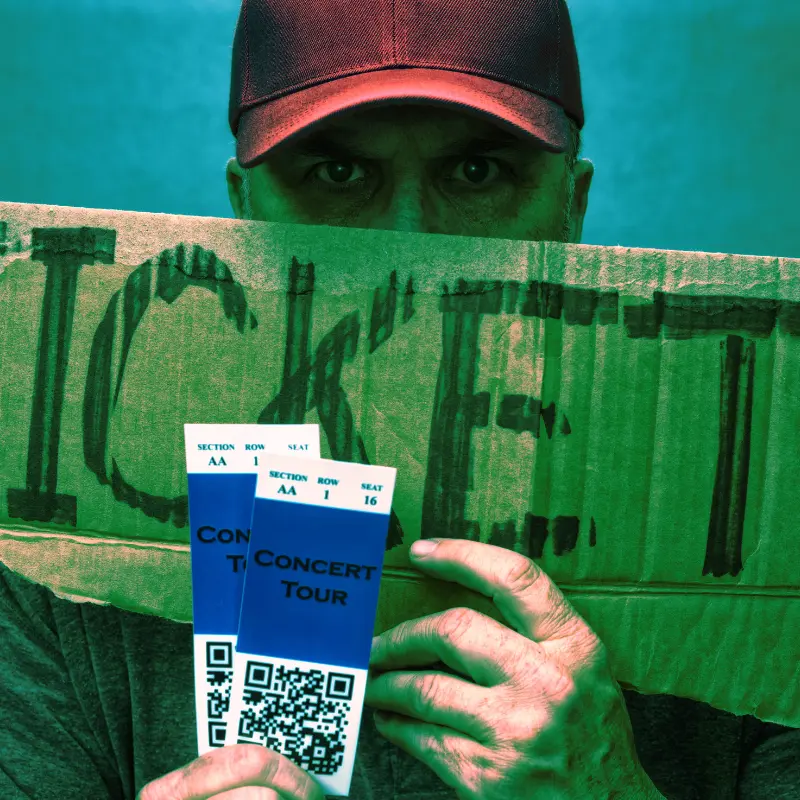 A man holding a cardboard sign reading tickets that covers most of his face and he's holding 2 concert tickets in front of the sign.