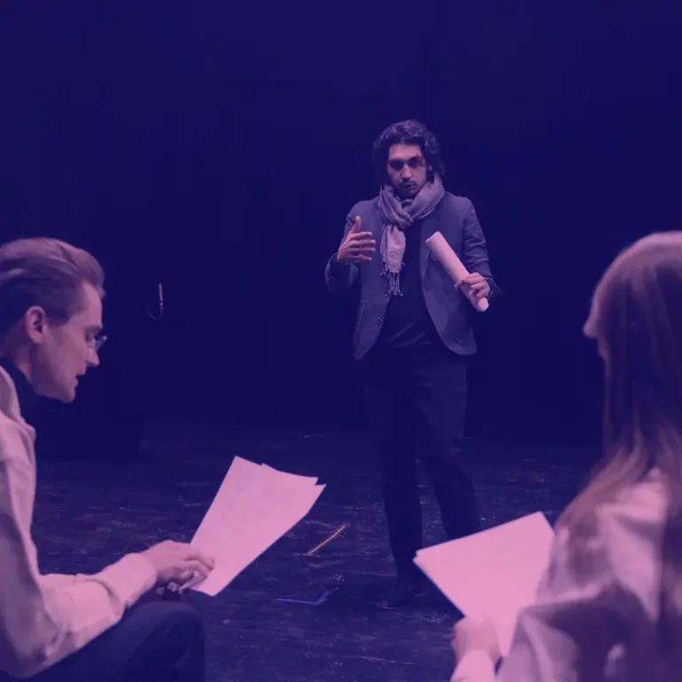 A man in a blazer and scarf passionately delivers a monologue during an acting audition on a dimly lit stage, facing two seated individuals with scripts.