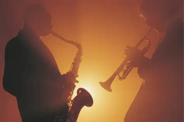 The warm glow of the image sets a moody atmosphere, highlighting two silhouetted jazz musicians, one with a saxophone and the other with a trumpet. They appear immersed in their performance, the soft backlighting creating an intimate vibe that's synonymous with a live jazz session, reminiscent of the experiences Jazz Live at Dizzy's offers to its audiences.