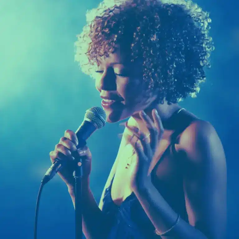 A female singer with curly hair performs passionately into a microphone, her expression conveying emotion, against a moody blue-lit background.
