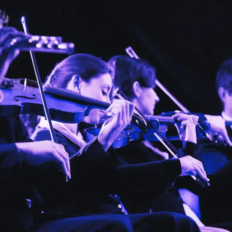A group of violinists in mid-performance during symphony auditions, illuminated by blue stage lighting