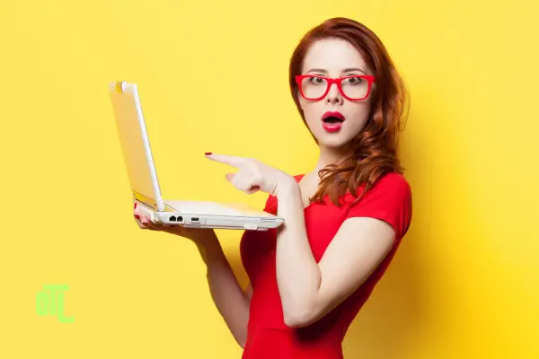 A woman in a red dress and red glasses expresses surprise while pointing to an open laptop she is holding, set against a bright yellow background.