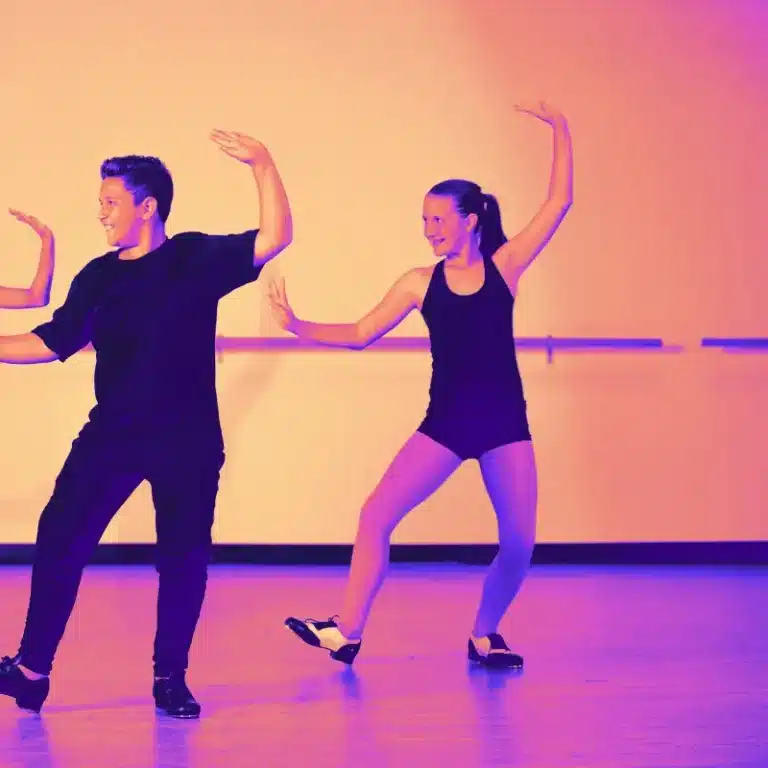 Two young dancers, a boy and a girl, in matching black outfits perform a tap dance routine on stage, with a warm-toned backdrop.