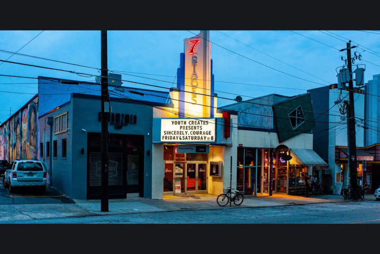 Exterior view of 7 Stages Theater in Atlanta at dusk, featuring its illuminated marquee promoting a youth production titled "Sincerely, Courage." The surrounding area includes adjacent buildings and street elements like power lines and parked cars.