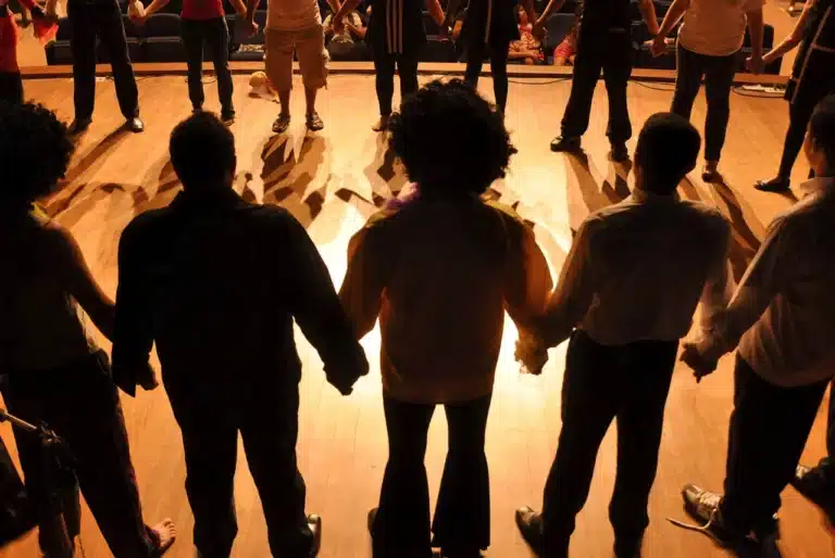 Performers holding hands on a theater stage, facing the audience under warm stage lighting. Shadows stretch across the wooden floor, capturing the essence of live theatre.