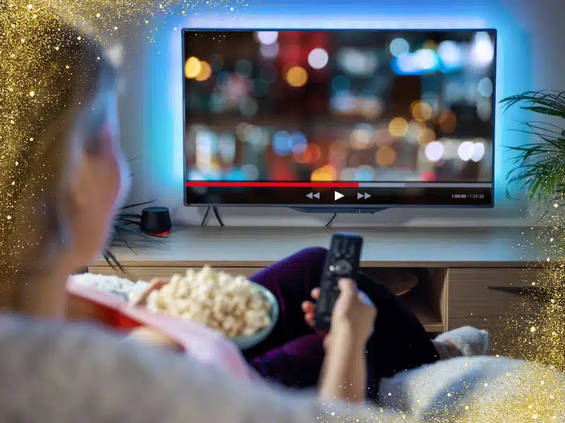 Person relaxing on a sofa with a bowl of popcorn, holding a remote while watching a TV streaming screen—evokes at-home performing arts streaming.