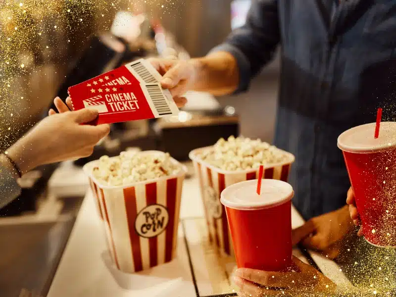 Close-up at a movie concession stand—someone hands over red cinema tickets beside striped popcorn tubs and red soda cups, with a festive gold-sparkle overlay.