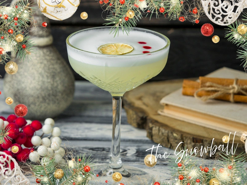 Snowball cocktail in a vintage coupe with a frothy top, lime wheel, and three red drops, framed by festive garlands and ornaments—an advocaat-and-lemonade classic perfect for UK panto season and holiday theatre nights.