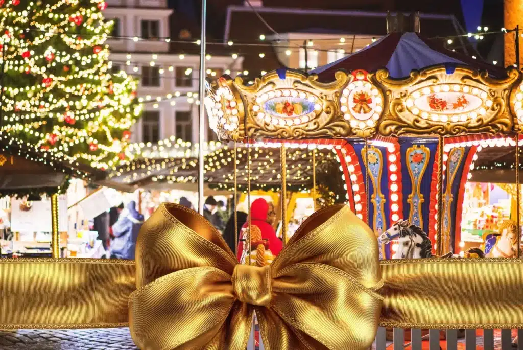 Festive holiday market with a brightly lit carousel, sparkling string lights, and a large gold ribbon in the foreground.