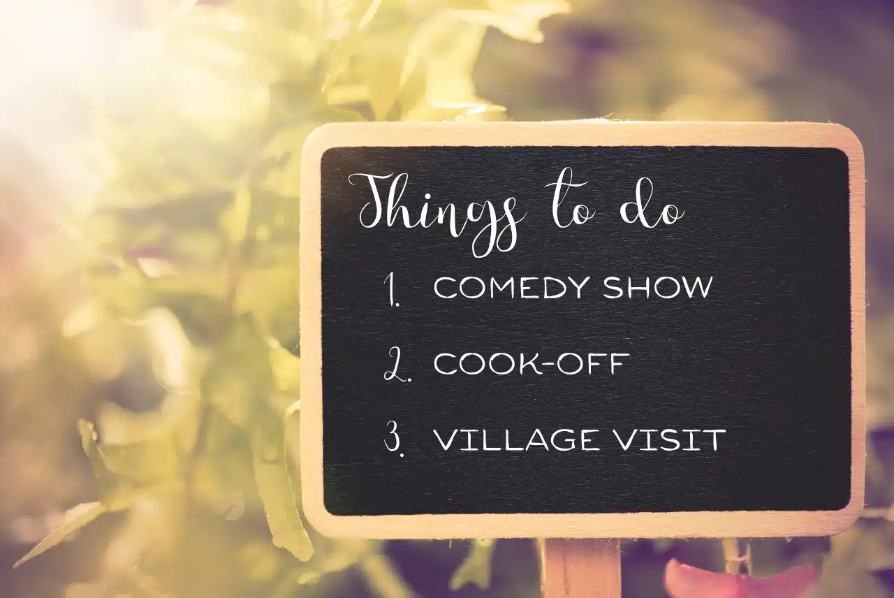Small chalkboard sign in a sunny garden listing “Things to do: 1. Comedy show, 2. Cook‑off, 3. Village visit.”