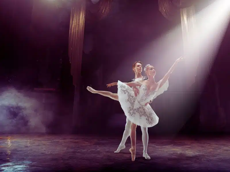 Ballet dancers performing a duet on stage in a spotlight with theatrical smoke and elegant curtains.