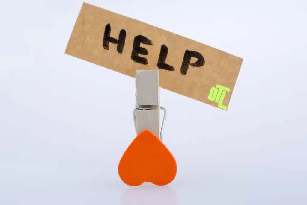 Clothespin holding a brown paper label that says “HELP,” with an orange heart clip and a small neon-green OTL logo on a white background.