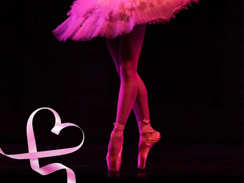 Ballet dancer en pointe under pink lighting with a ribbon shaped like a heart on the stage floor.