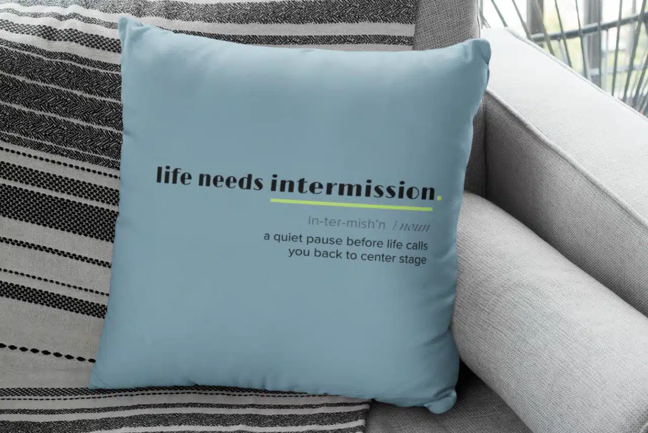 Life Needs Intermission Busy Life Edition light blue throw pillow on a gray couch.
