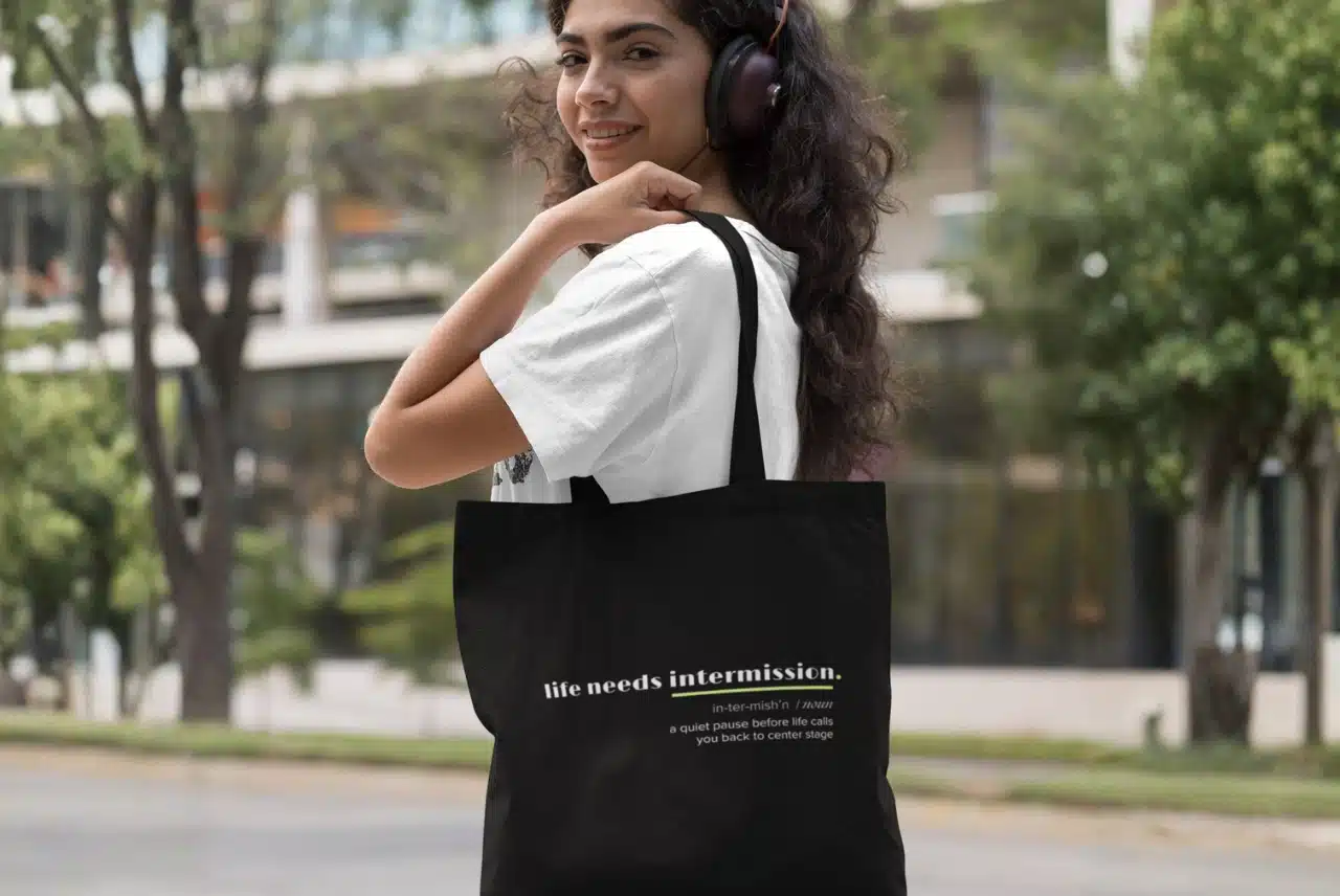 Life Needs Intermission Busy Life Edition black tote bag worn over the shoulder outdoors.