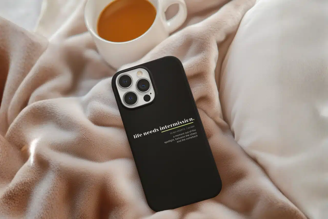 Life Needs Intermission Chaos Edition black phone case on a soft blanket with a mug nearby.