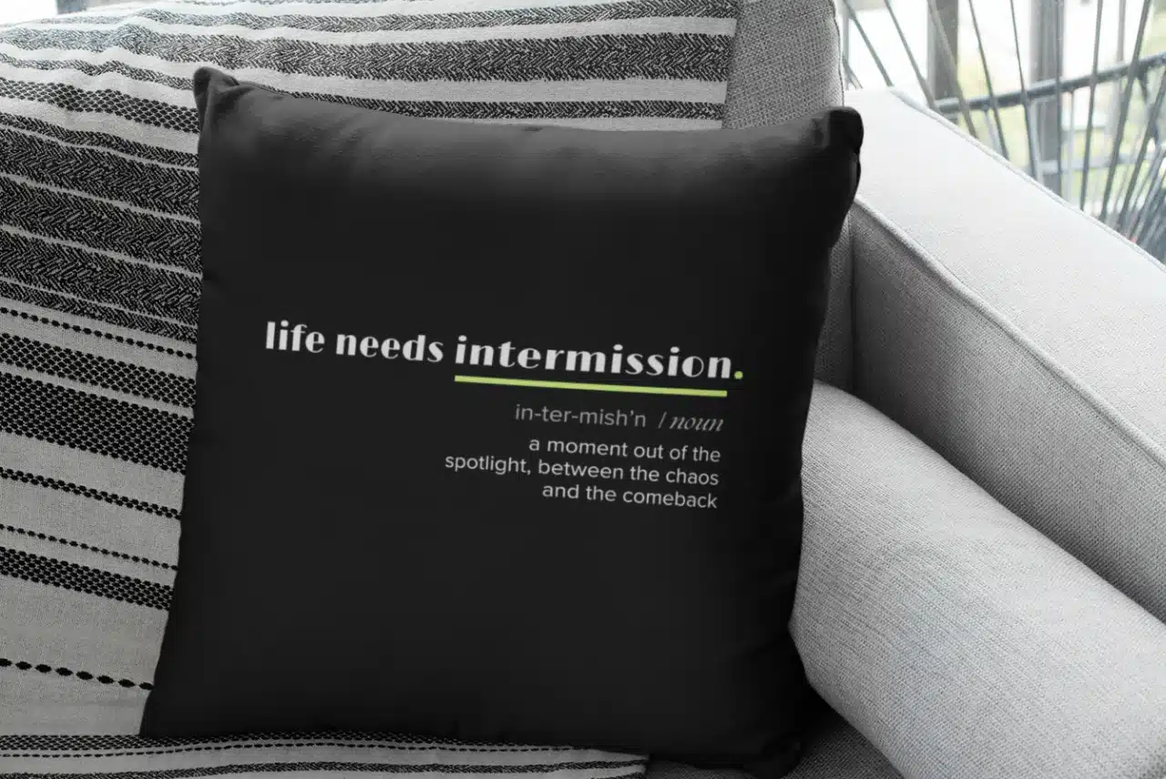 Life Needs Intermission Chaos Edition black throw pillow with definition-style text on a gray couch.