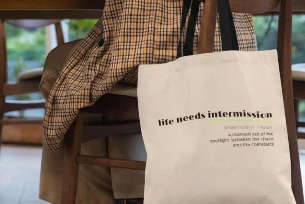 Life Needs Intermission Chaos Edition cream tote bag hanging from a wooden chair.