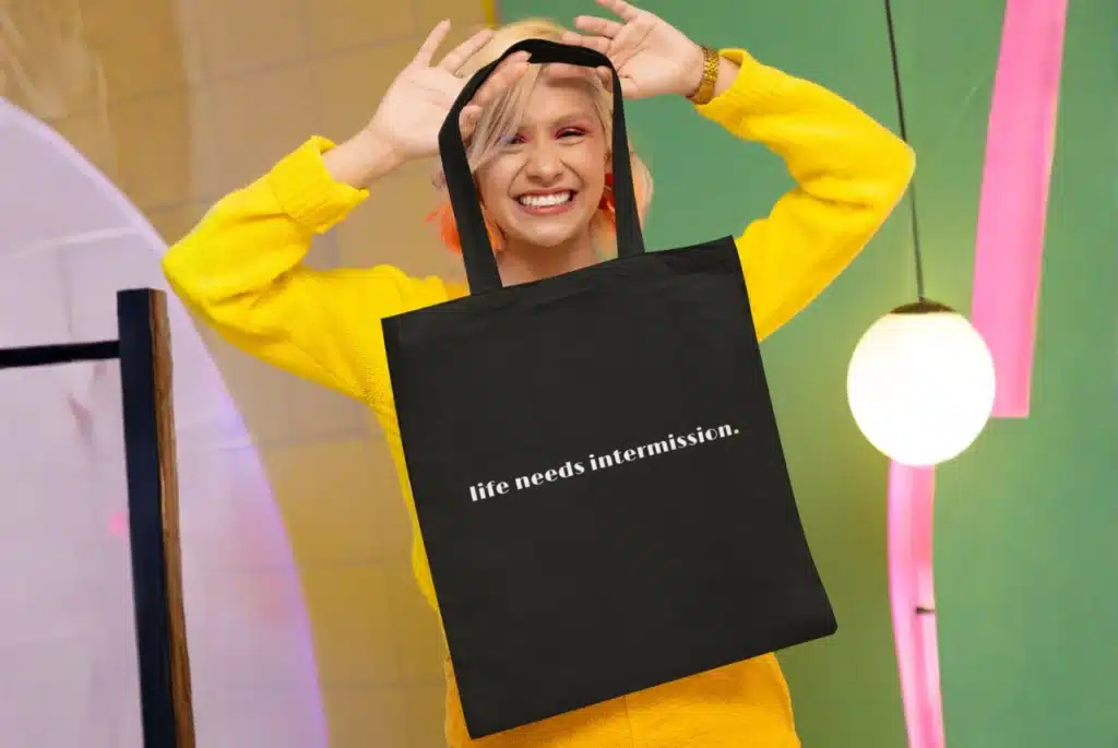 Smiling woman in a bright yellow sweater holding a black tote bag printed with the phrase “life needs intermission.” against a colorful modern backdrop.
