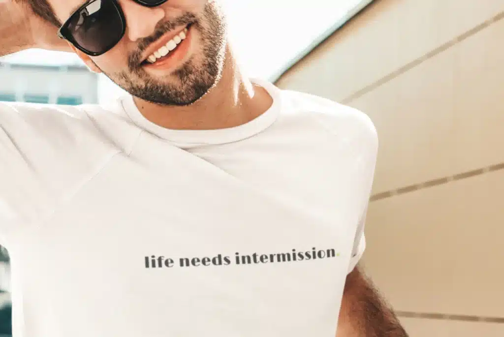 Man modeling a white “life needs intermission.” t-shirt with dark text and a small green period.