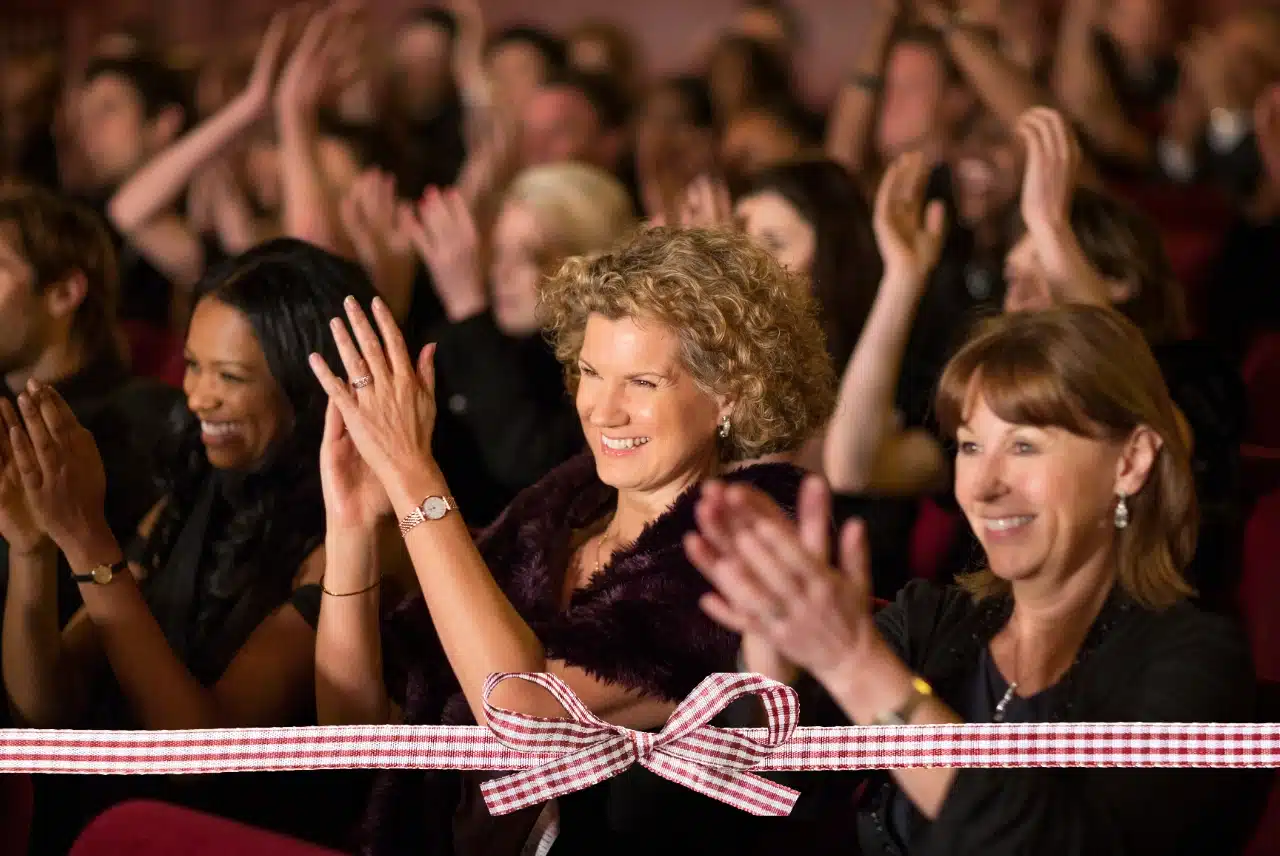 Three smiling women applauding at a live theater performance with a red-and-white gift ribbon across the bottom
