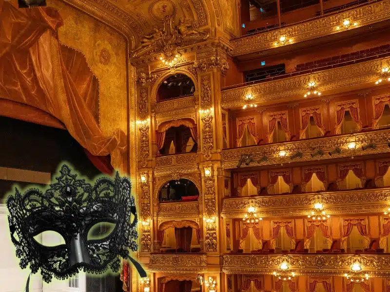 Ornate gold opera house interior with tiered balconies, red drapery, and a black lace masquerade mask in the foreground.