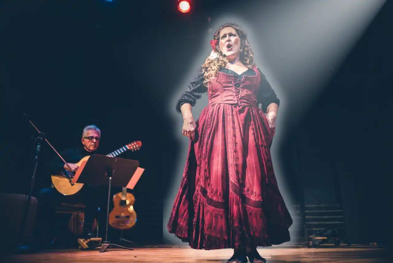 Opera singer in a deep red gown performing on stage under a bright spotlight, with a seated guitarist accompanying her in the background.