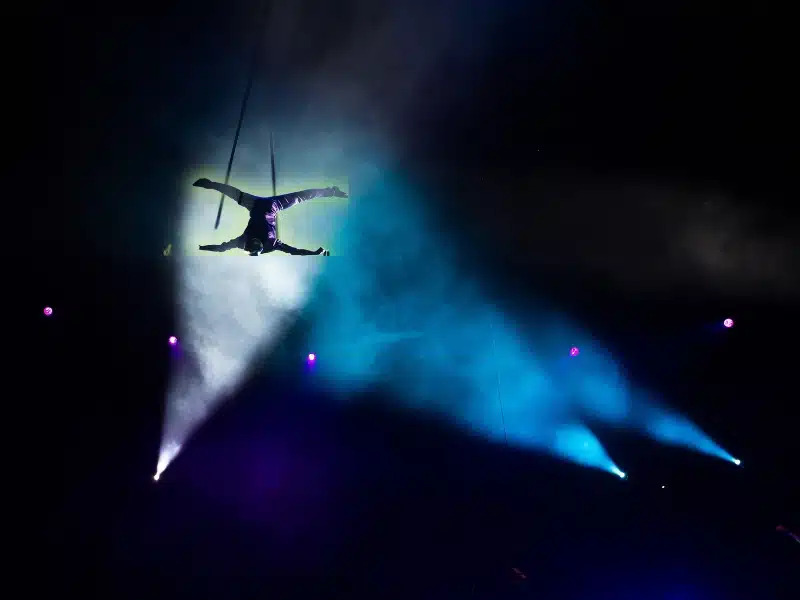 Aerial circus performer suspended midair under colorful stage lighting