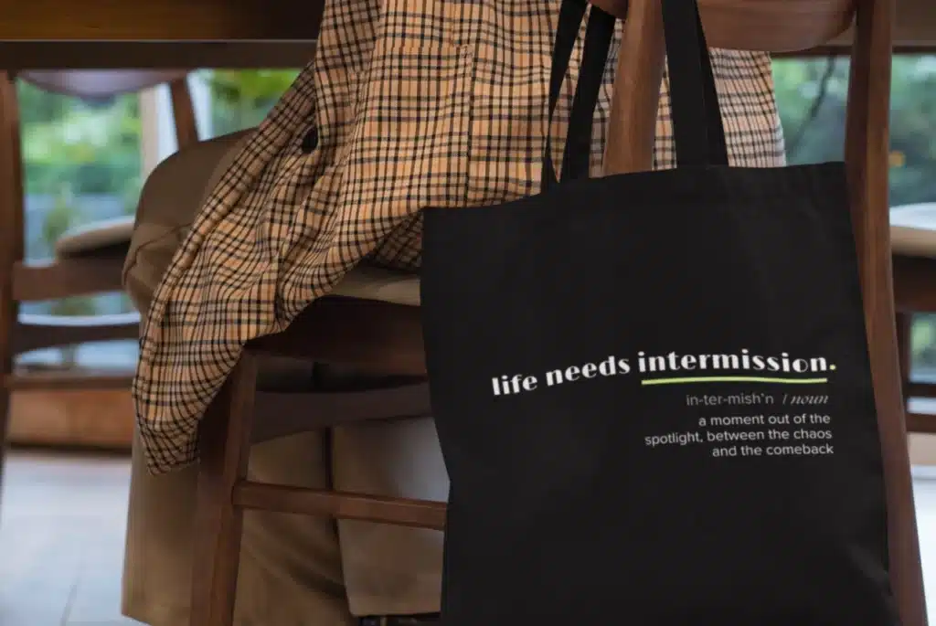 Life Needs Intermission Chaos Edition black tote bag hanging over the back of a chair.