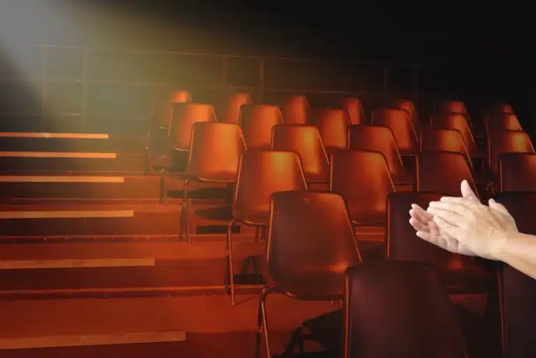 Fringe theatre audience seats lit by warm stage lights, with hands applauding beside an intimate performance space.