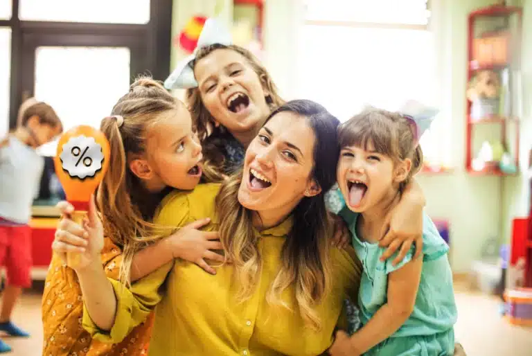 Teacher smiling with three children in a classroom while one child holds a colorful discount sign.