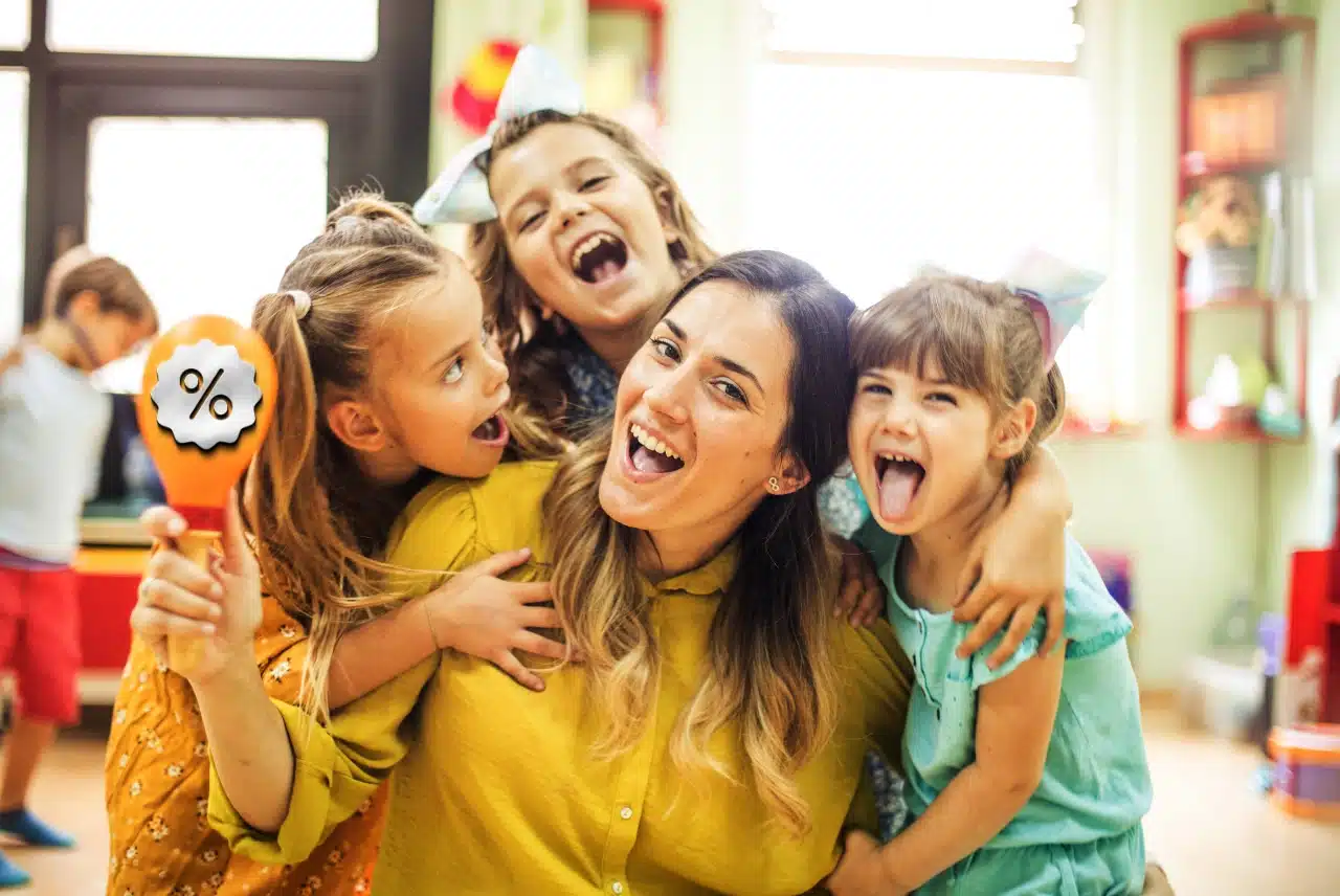 Teacher smiling with three children in a classroom while one child holds a colorful discount sign.