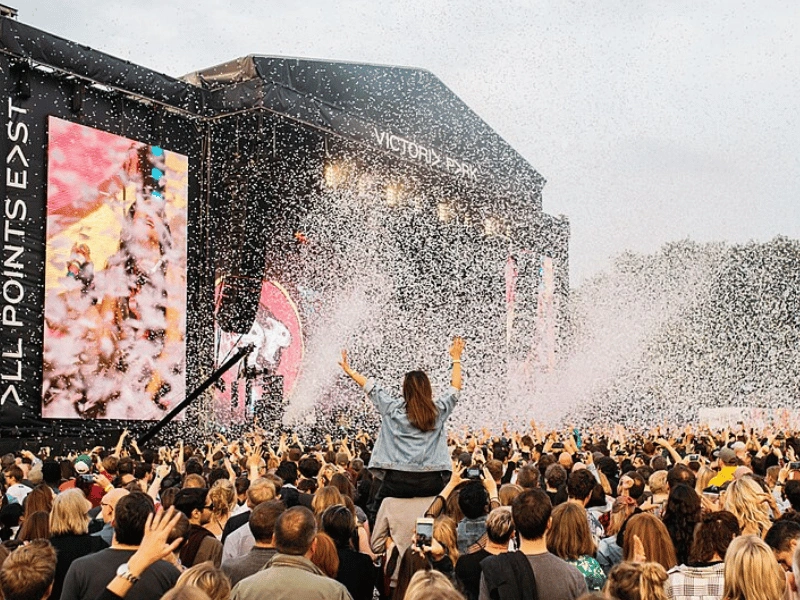 A large crowd celebrating beneath a burst of confetti in front of the All Points East stage at Victoria Park.