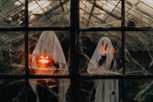 Two people in white sheet ghost costumes standing behind cobweb-covered greenhouse windows at dusk, one holding a glowing carved jack-o’-lantern and the other looking at a mobile phone.