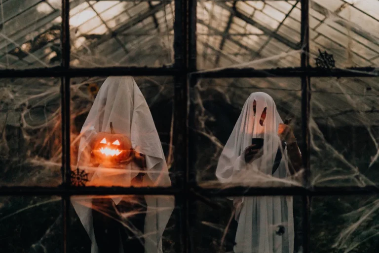 Two people in white sheet ghost costumes standing behind cobweb-covered greenhouse windows at dusk, one holding a glowing carved jack-o’-lantern and the other looking at a mobile phone.